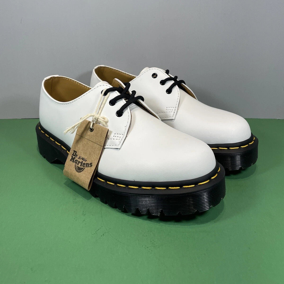 A pair of white Dr. Martens 163 Classic shoes with black laces, black soles, and a brown paper brand tag, displayed on a green surface against a gray background.