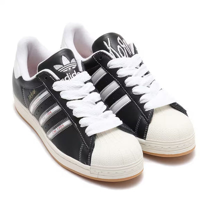 Adidas Superstar x Korn Est. 1994 collaboration sneakers in black leather with silver stripes, off-white shell toe, chunky white laces, and gum sole — Swingdot Luxe