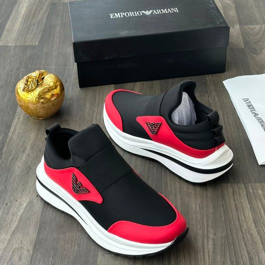 A pair of Emporio Armani slip-on sneakers in black and red, displayed on a gray wood floor alongside their black shoebox, a small gold decorative object, and a paper card.