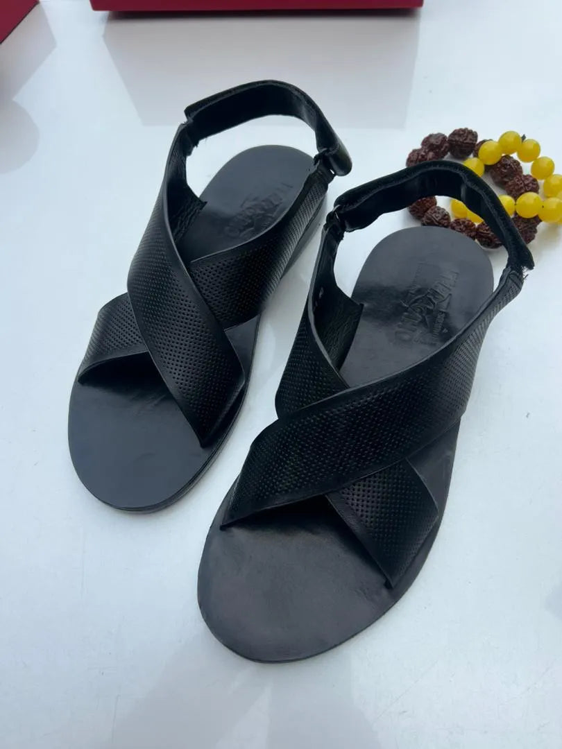 A pair of black leather criss-cross strappy sandals with adjustable ankle straps, displayed on a white surface next to a yellow beaded bracelet.
