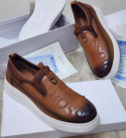 A pair of brown leather slip-on platform sneakers with 'BOSS' embossed on the toe and side, displayed on top of their white shoeboxes with paperwork.