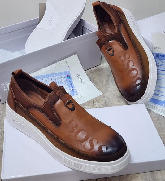 A pair of brown leather slip-on platform sneakers with 'BOSS' embossed on the toe and side, displayed on top of their white shoeboxes with paperwork.