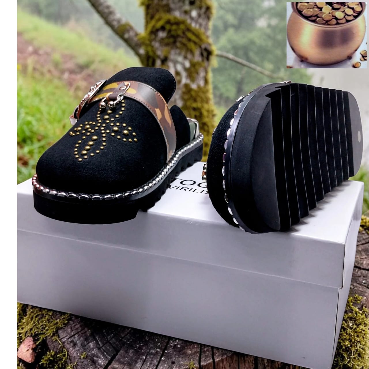 Black suede loafer mule with gold stud motif, camo-print gold chain strap, crystal perimeter trim, and chunky ridged black rubber sole — displayed on a white box in a natural outdoor setting