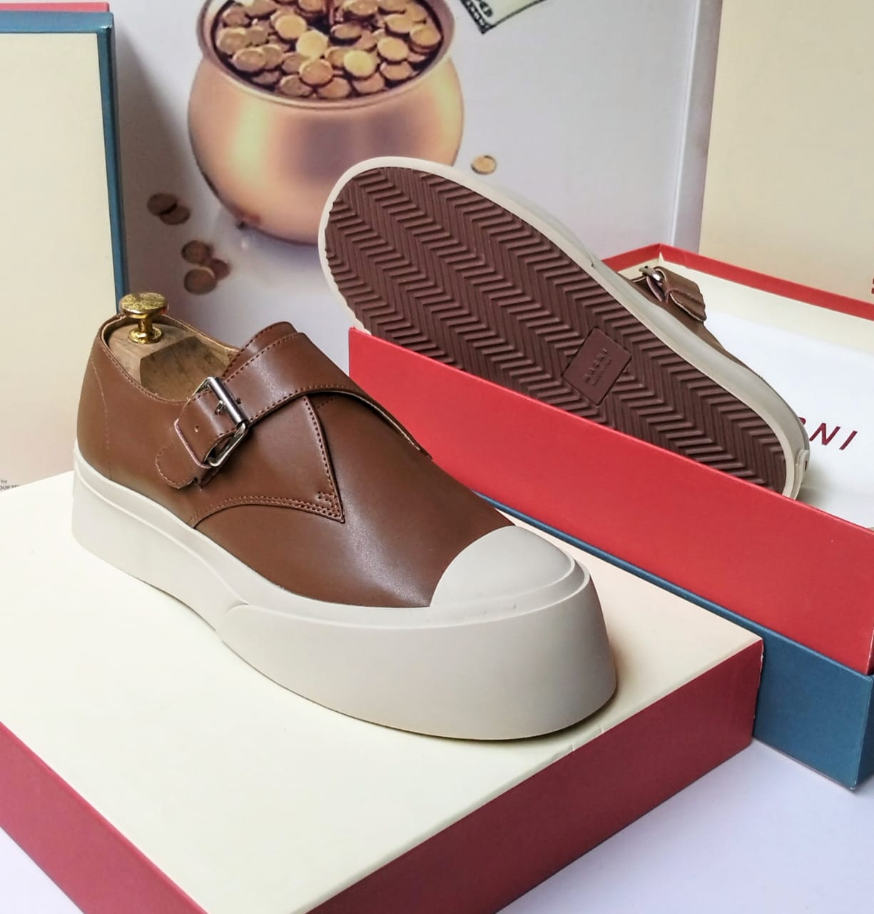 Tan calfskin leather monk strap platform sneaker with polished silver buckle, white cap toe, and thick white herringbone rubber sole — side and sole view on a red luxury shoe box