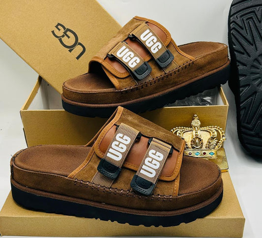 UGG Tasman X chestnut suede double-strap slides with bold UGG wordmark webbing straps in tan and white, black buckle hardware, espadrille midsole stitching, and black ridged rubber outsole on UGG kraft box — 100% authentic men's luxury slides