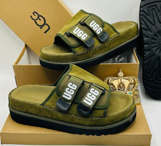 UGG Tasman X olive moss green suede double-strap slides with tonal UGG wordmark webbing straps in dark green and white, black buckle hardware, espadrille midsole stitching, and black ridged rubber outsole on UGG kraft box — 100% authentic men's luxury slides
