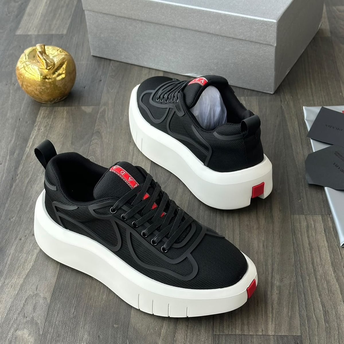A pair of black platform sneakers with thick white soles and small red accents, displayed on a dark wood floor next to a gray shoebox, a gold decorative object, and some papers.