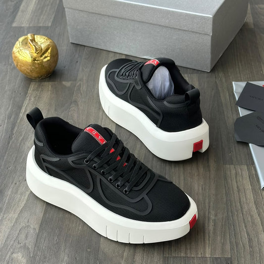 A pair of black platform sneakers with thick white soles and small red accents, displayed on a dark wood floor next to a gray shoebox, a gold decorative object, and some papers.